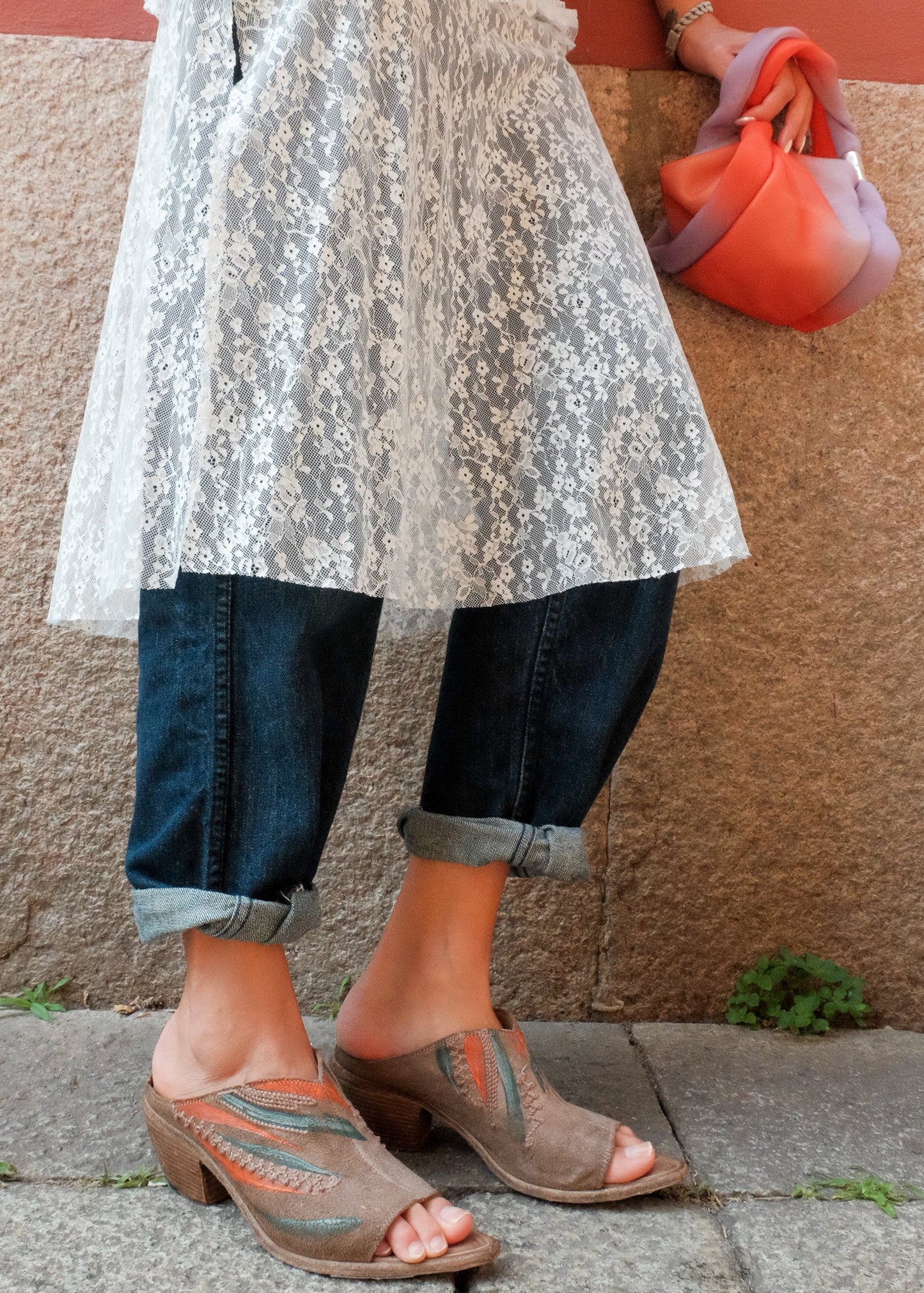 Person wearing a white lace dress, blue jeans, and brown sandals against a textured wall.