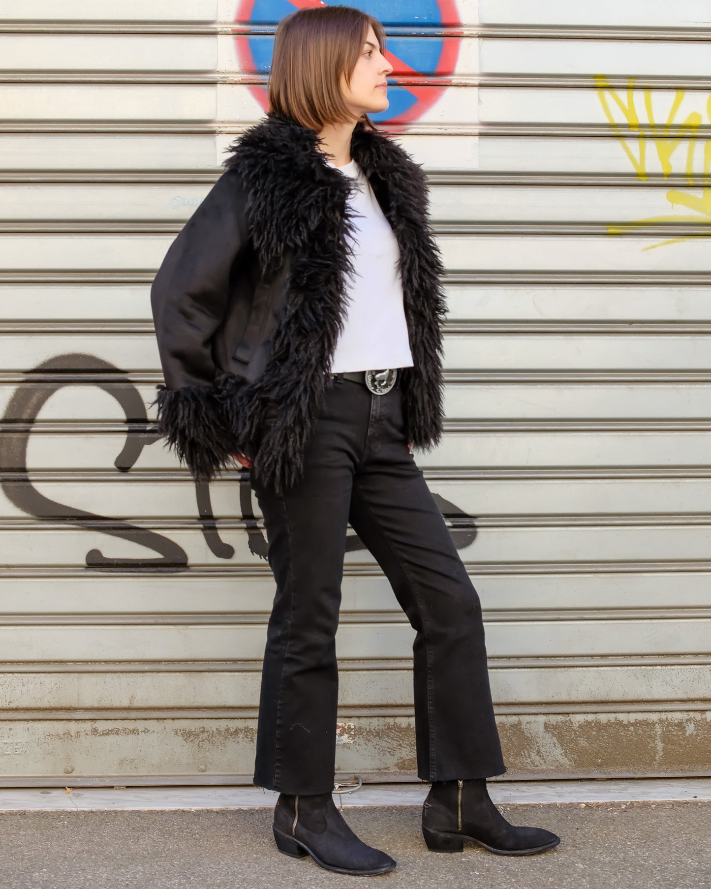 Person wearing a black fur coat, white shirt, black pants, and black boots standing in front of a graffiti-covered wall.